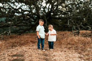 Family Photographer, two young boys stand together happy in dry grass beneath the trees