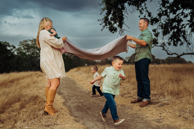 Family Photographer, a mother and father hold a blanket up as their young children run beneath it outside
