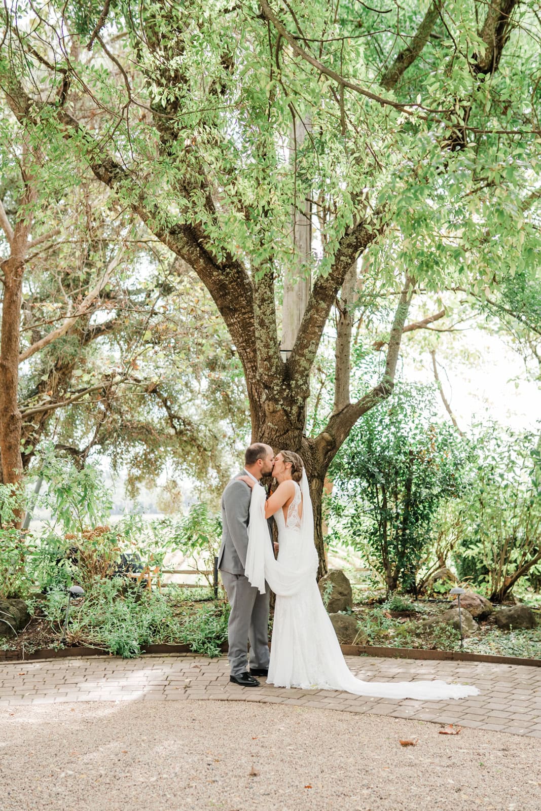 A bride and groom kiss on a garden path under trees
