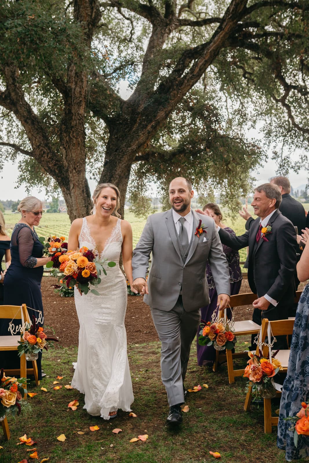Newlyweds hold hands and happily exit their outdoor ceremony up the aisle under a large tree as guests celebrate at one of the Sonoma wedding venues