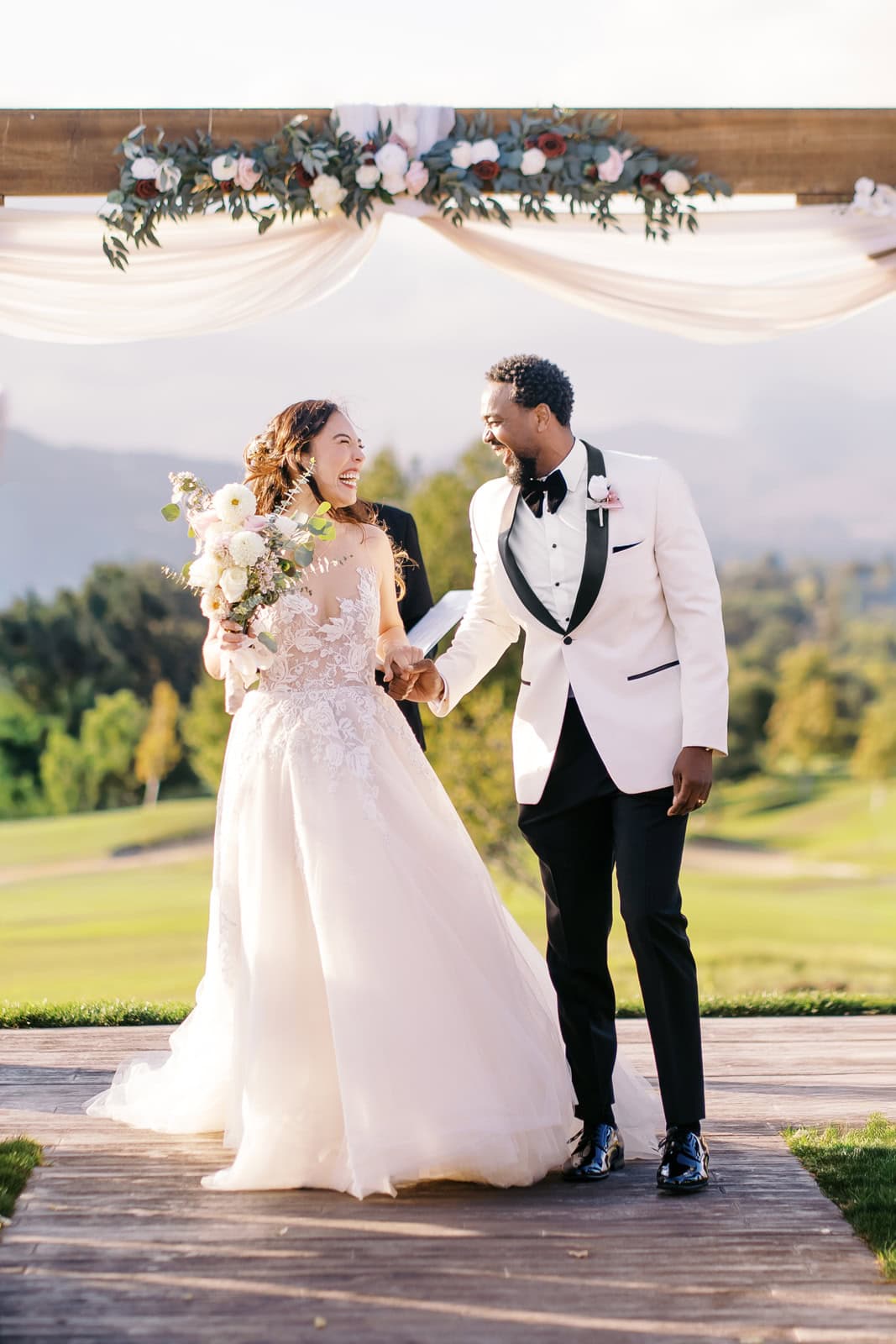 Laughing newlyweds hold hands while smiling at each other and walking up the aisle at sunset to end their outdoor ceremony at one of the wedding venues in Brentwood, CA