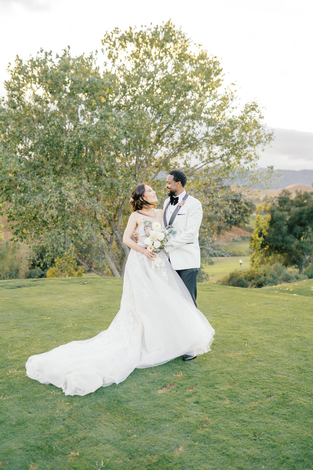 Happy newlyweds smile while hugging in a white tux jacket in a hilltop lawn at sunset at one of the wedding venues in Brentwood, CA